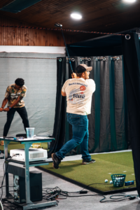Two individuals swing golf clubs at an indoor simulator with artificial turf and tracking equipment.