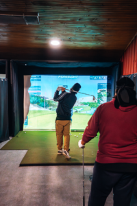 A golfer swings on an indoor simulator hitting mat, while another watches. The screen displays a virtual golf course landscape.