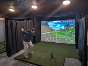 A golfer practices indoors in a virtual golf bay. A large screen displays a virtual golf course with trees and sky.