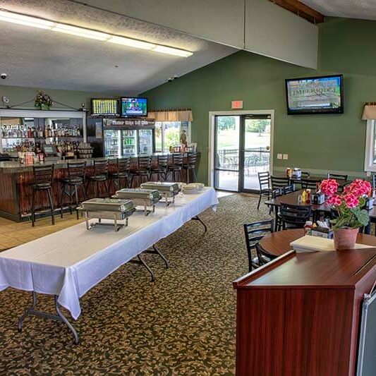 A restaurant with green walls, a bar area on the left, and a long table with a white tablecloth set up for a buffet. Tables and chairs are arranged on a patterned carpet, and a potted plant sits on a podium.