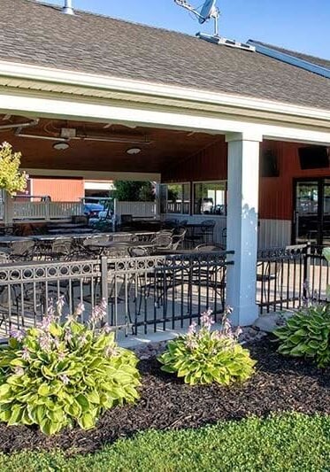 A Covered Outdoor Patio With Black Metal Tables And Chairs, Surrounded By Lush Landscaping Including Flowering Bushes And Green Plants, Under A Clear Blue Sky A covered outdoor patio with black metal tables and chairs, surrounded by lush landscaping including flowering bushes and green plants, under a clear blue sky.