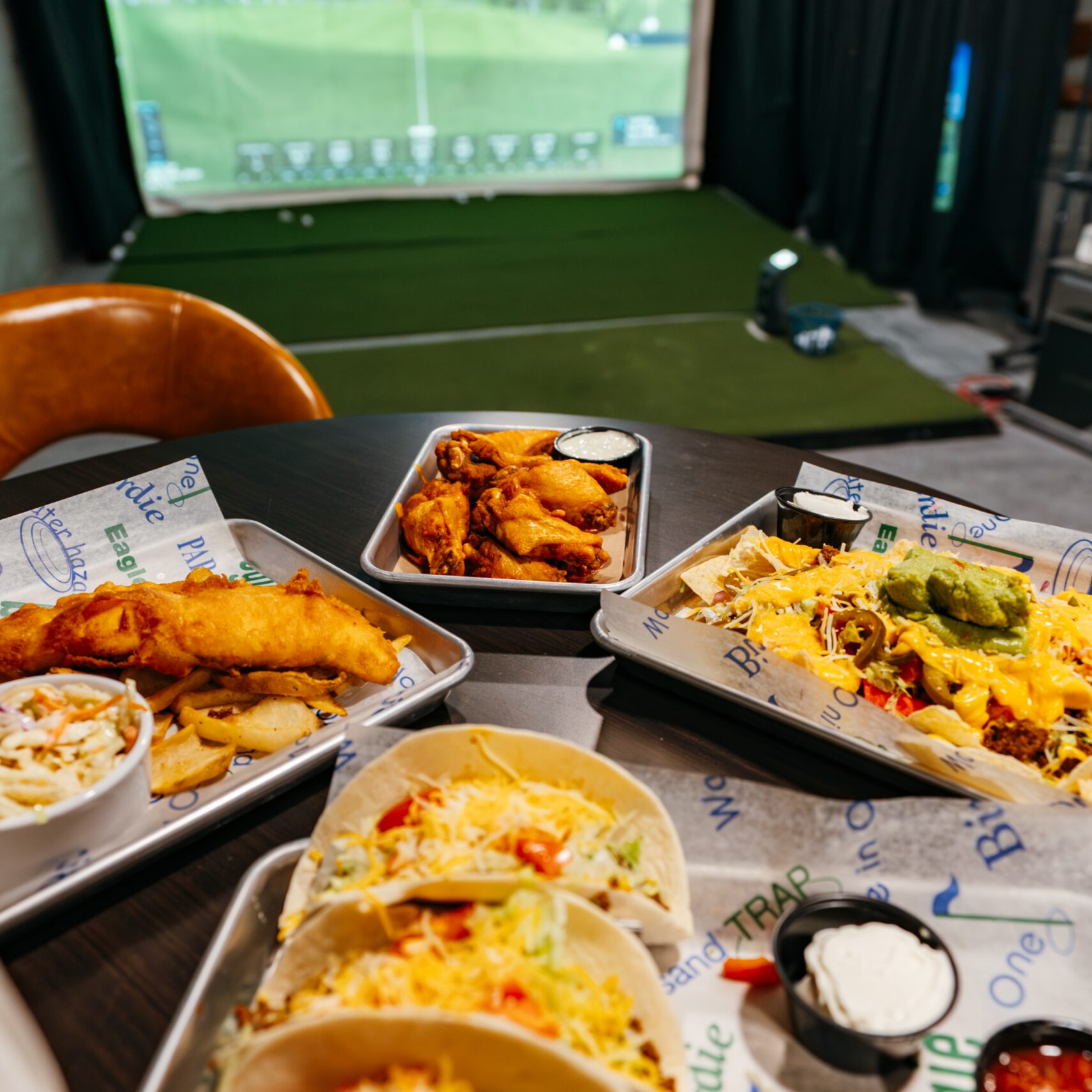 A table with tacos, fish, and chips in the foreground and an indoor golf simulator screen in the background. The foil-lined food containers are placed on a dark tabletop near a golf mat.